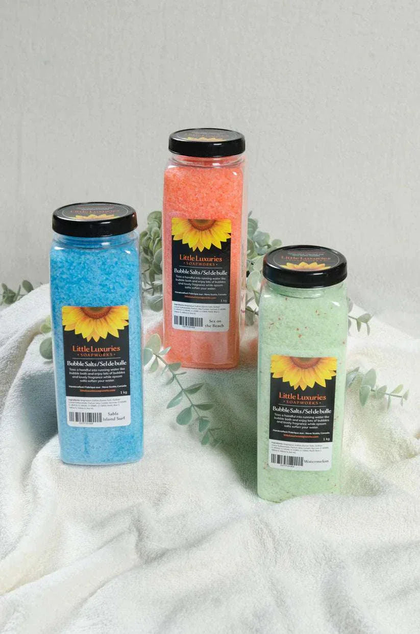 Three colorful bottles of bubble bath salts in blue, orange, and green with black lids on a soft white towel background.