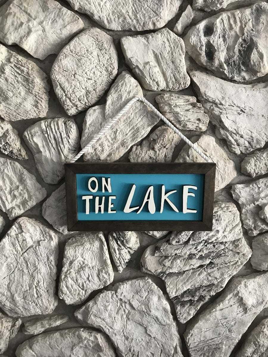 Handcrafted wooden sign with hand-drawn letters "On The Lake" in white on blue background with dark wood frame and rope hanger against stone wall