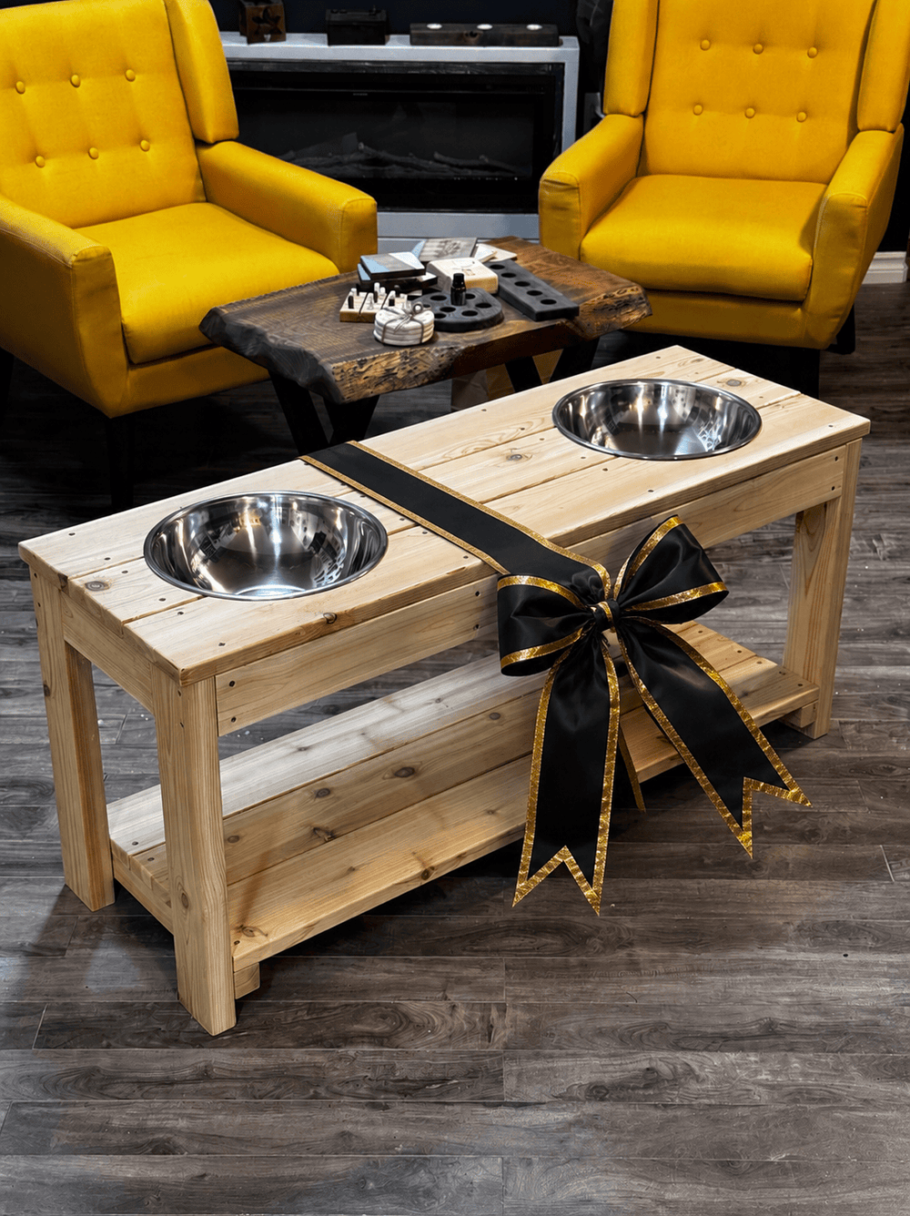Wooden pet stand with two elevated metal bowls, decorated with a black bow, in a room with yellow chairs.
