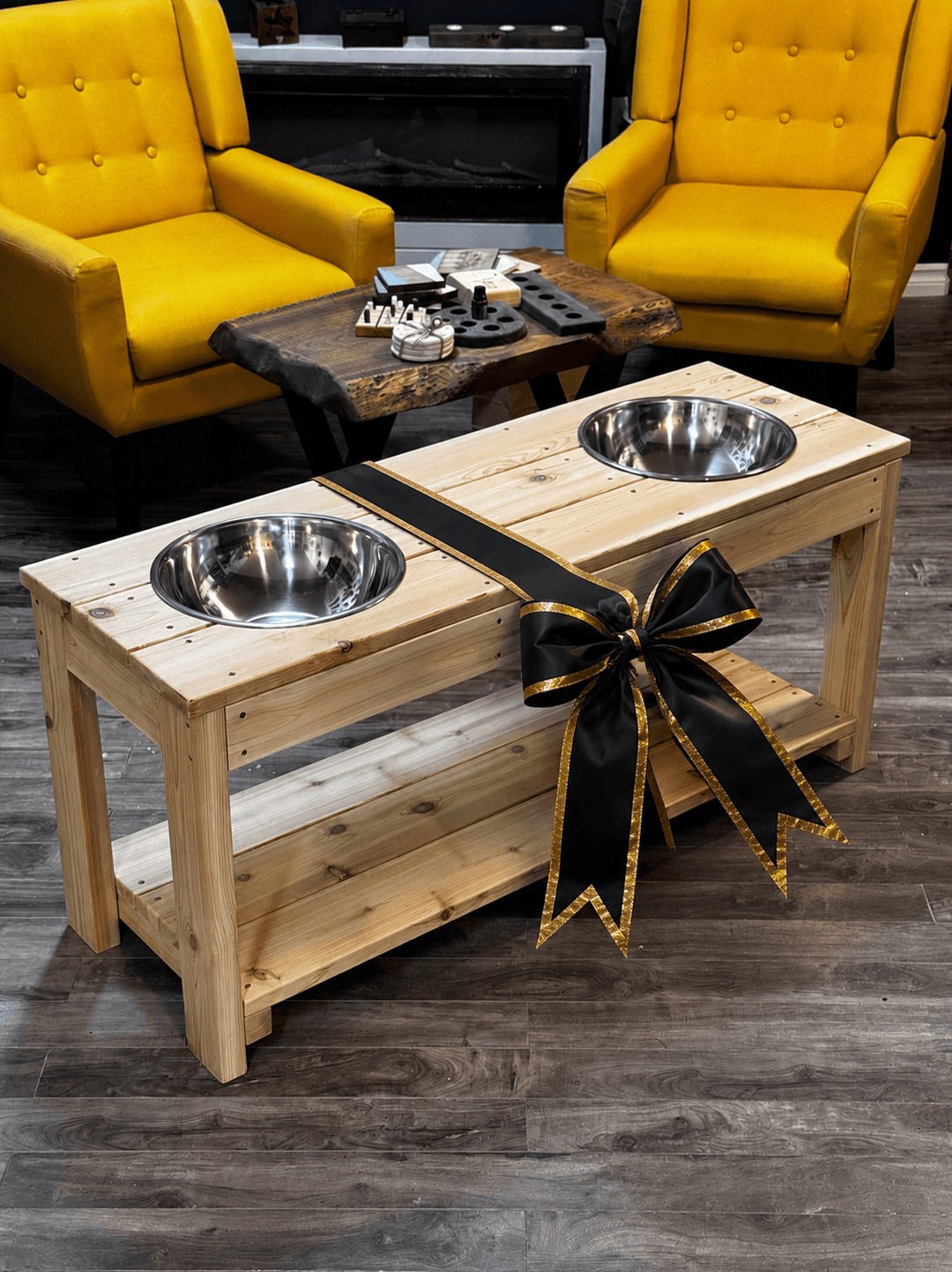 Wooden pet stand with two elevated metal bowls, decorated with a black bow, in a room with yellow chairs.
