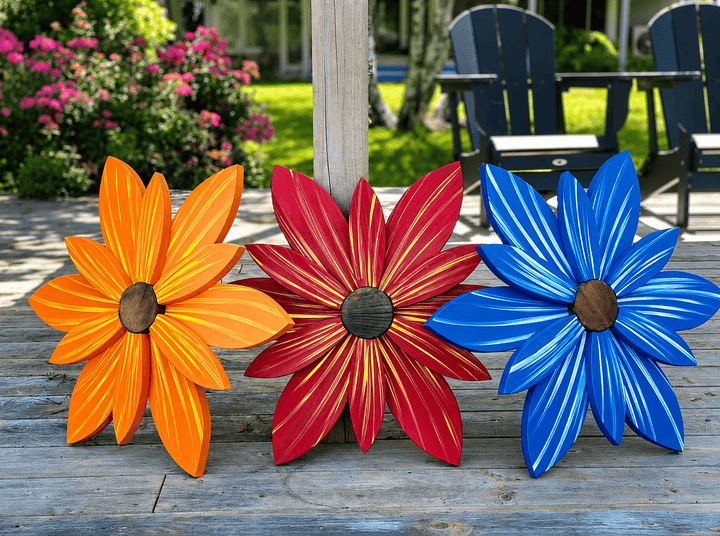 Three colorful metal flower sculptures in orange, red, and blue on a wooden surface with a garden background.