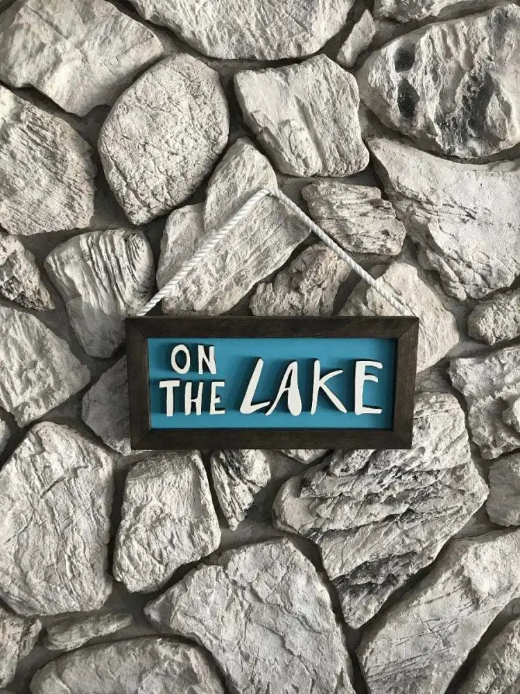 Handcrafted wooden sign with hand-drawn letters "On The Lake" in white on blue background with dark wood frame and rope hanger against stone wall