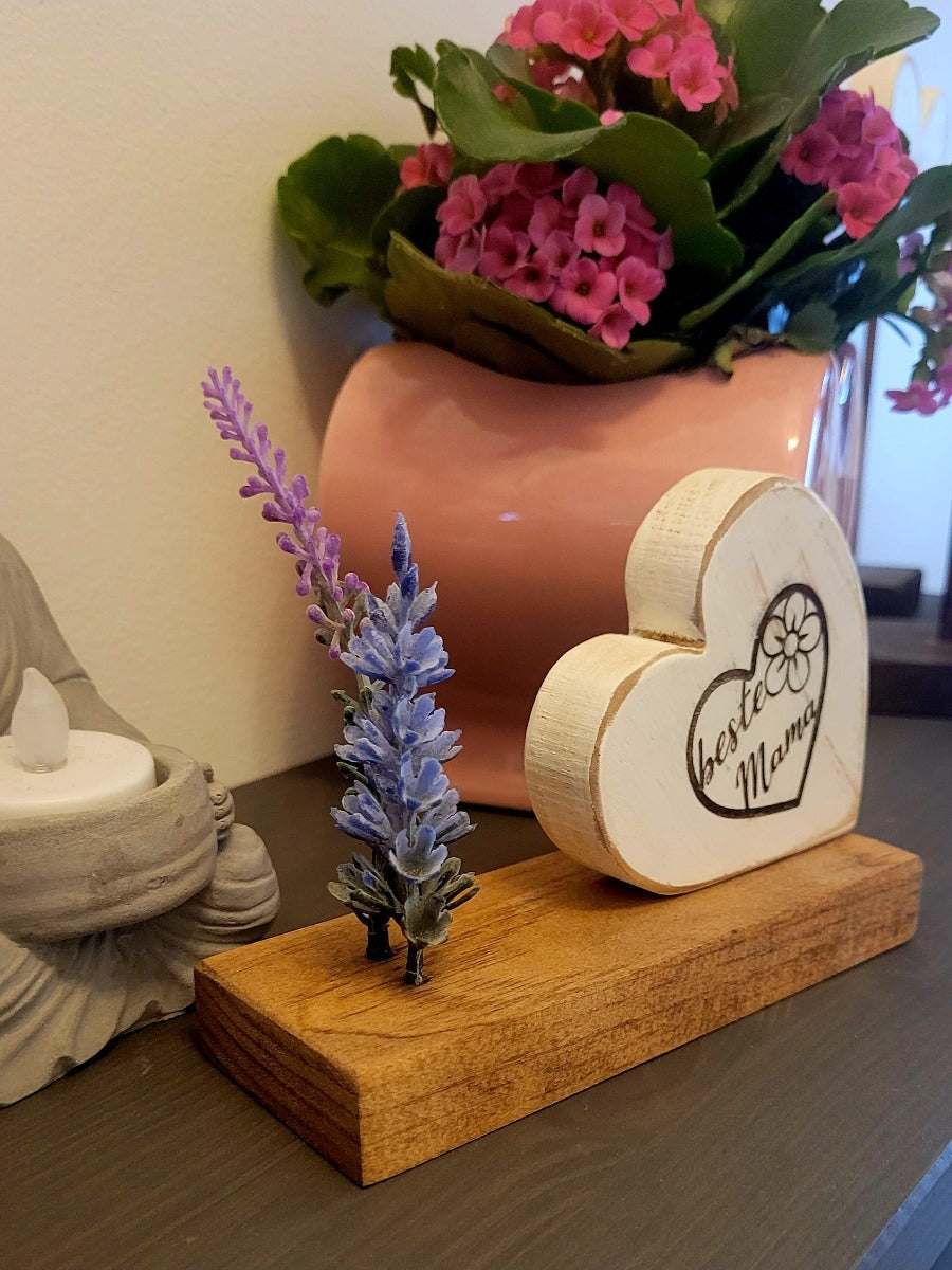 Wooden heart decoration "Beste Mama" on wooden base with lavender sprig, pink flowers in the background.