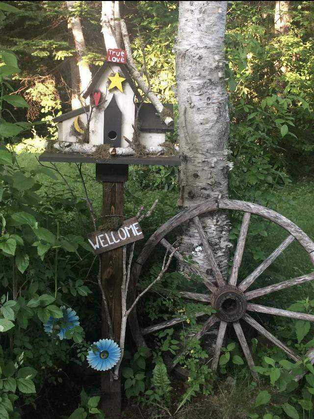 Triple birdhouse in garden with rustic design, featuring three compartments, personalized decorations, and surrounded by garden decor and vegetation.