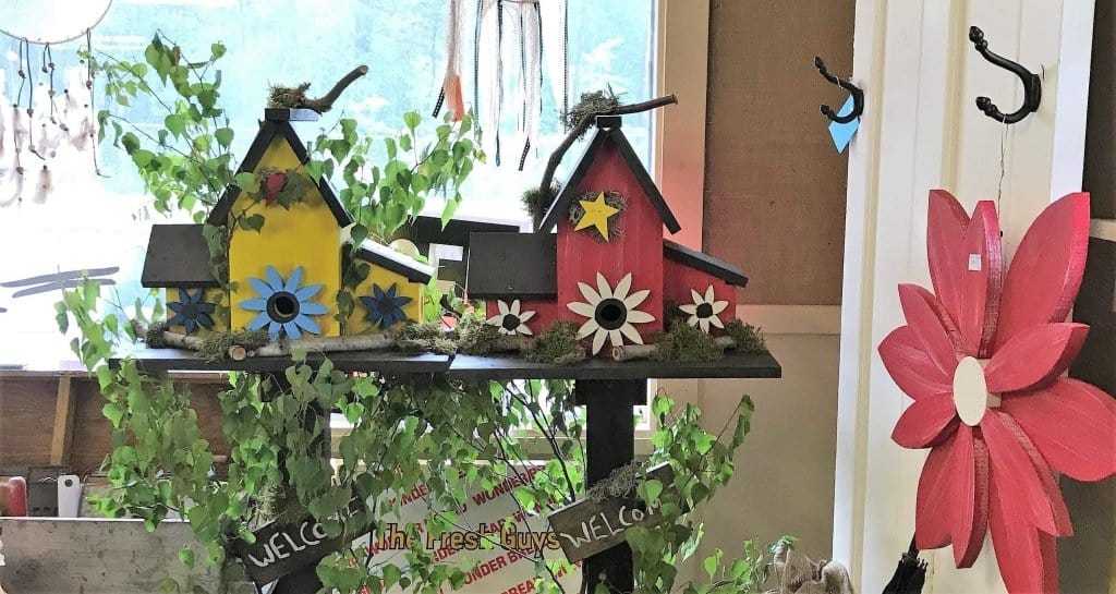 Colorful triple wooden birdhouses with flower decorations displayed indoors on a shelf surrounded by greenery and a large red flower decoration nearby