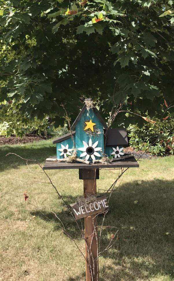 Triple birdhouse with three compartments, perched on a stand, featuring custom colors and designs in a garden setting.