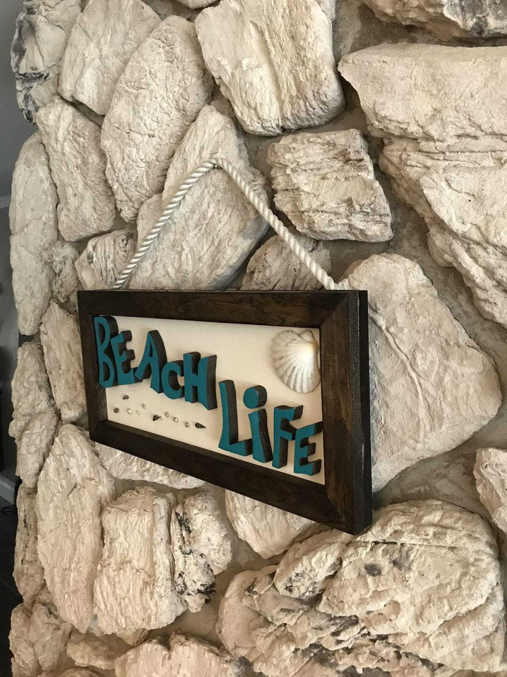 Hand painted wooden sign with "Beach Life" text, rope attachment, 24x11.5 inches, on textured stone wall.