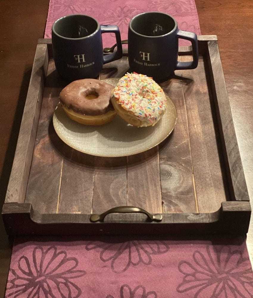 Antiqued rustic pine wood serving tray with metal handles holding two coffee mugs and a plate of donuts on a wooden table