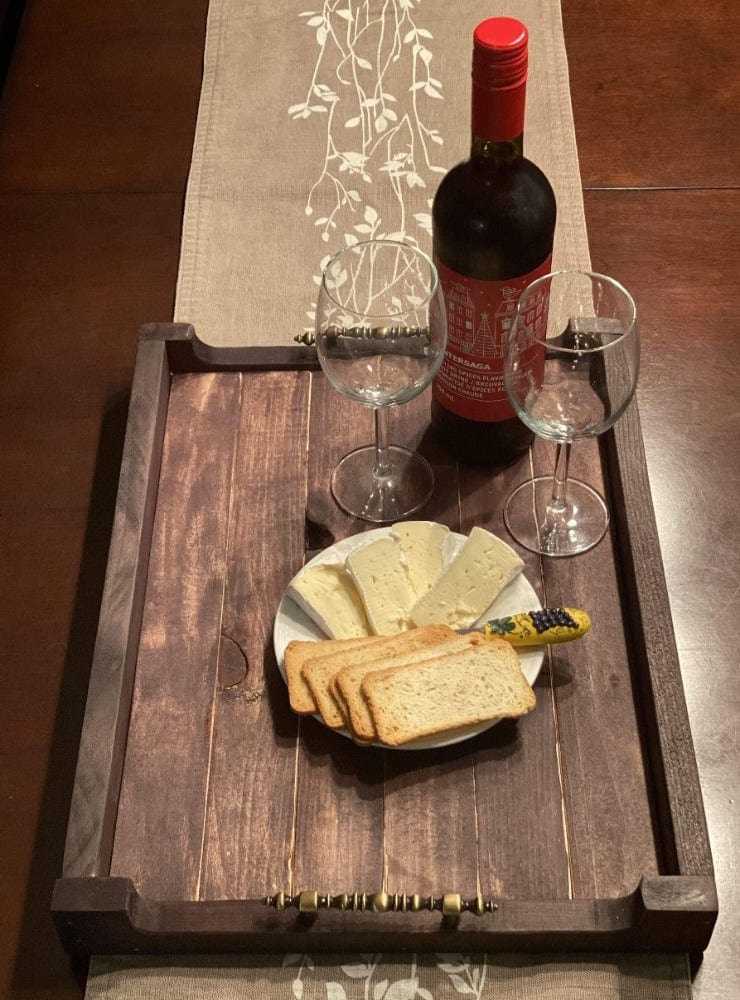 Handcrafted antiqued rustic pine wood serving tray with decorative metal handles, holding a wine bottle, two empty wine glasses, and a plate with cheese and crackers on a table runner.
