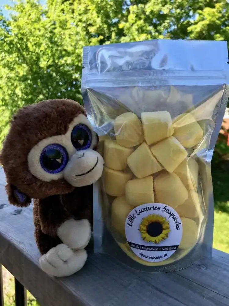 Clear resealable pouch of yellow bath bombs with sunflower label next to a plush monkey toy on outdoor wooden railing