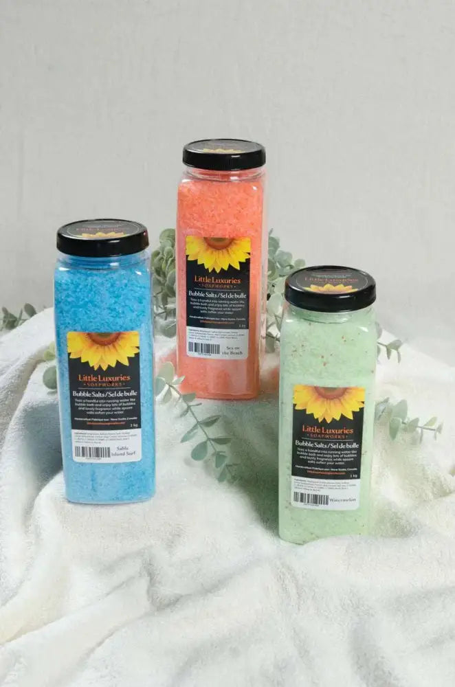 Three colorful bottles of bubble bath salts in blue, orange, and green with black lids on a soft white towel background.