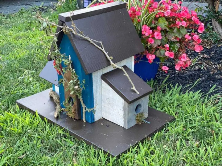Triple birdhouse with round entrances and perches, made of pine wood, in garden setting with pink flowers.