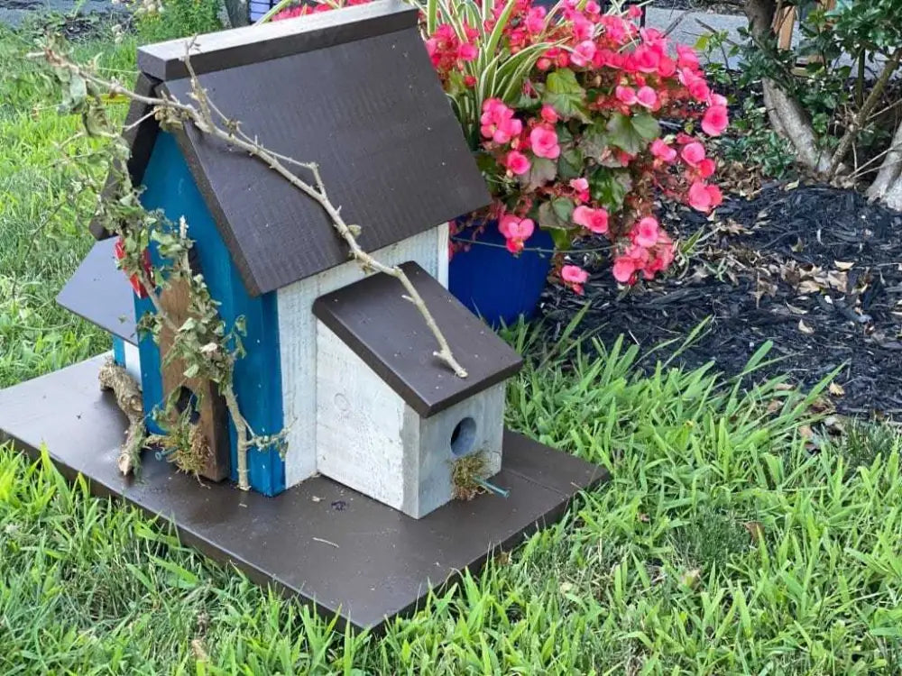 Triple birdhouse with brown roof and branches, set in a garden next to a pot of vibrant pink flowers.