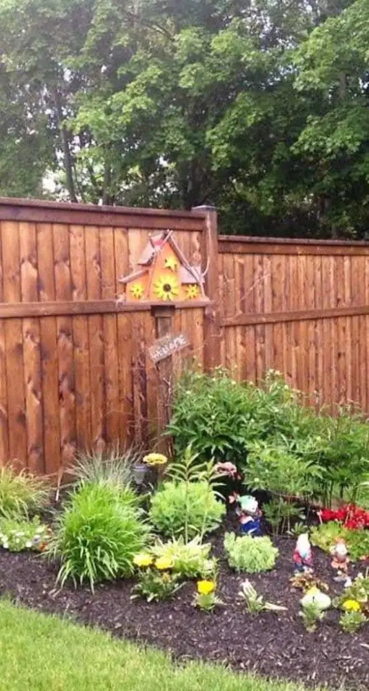 Triple pine wood birdhouse with three compartments mounted on wooden fence above garden bed with green plants and flowers