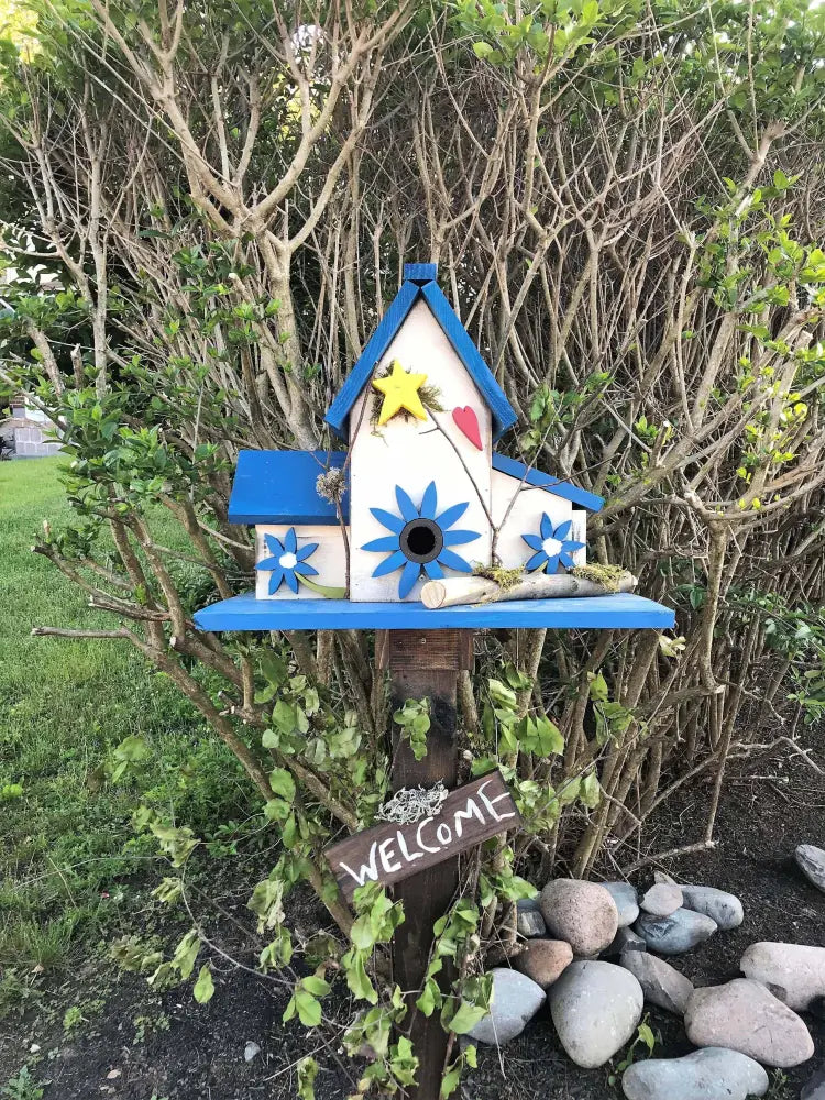 Triple birdhouse with blue roof, flower decorations, and "Welcome" sign mounted on a wooden post in a garden setting
