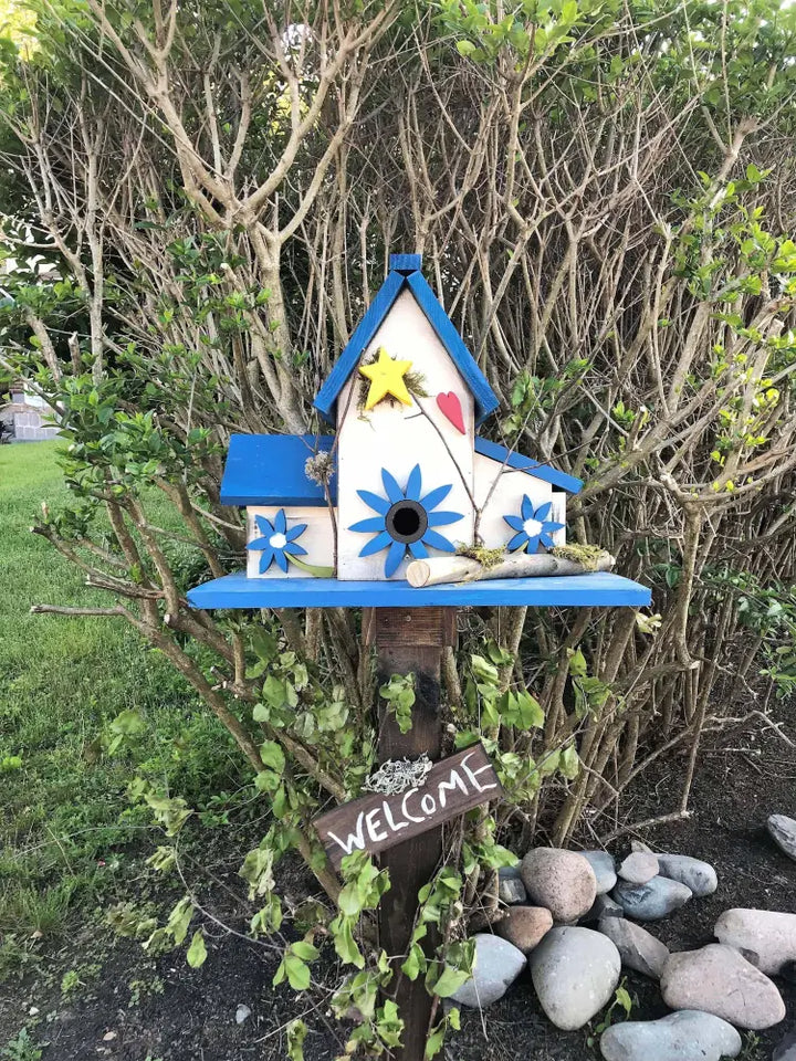 Triple birdhouse with blue roof, flower decorations, and "Welcome" sign mounted on a wooden post in a garden setting