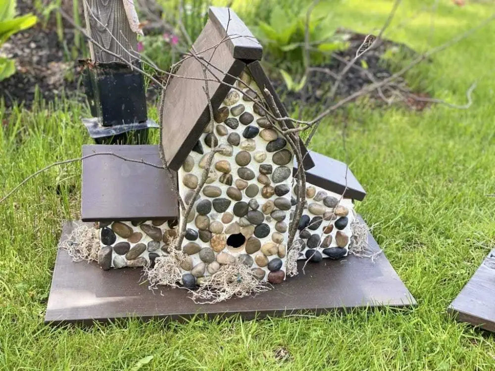 Triple birdhouse made from pine wood with stone pebble design, featuring three compartments, in a garden setting.