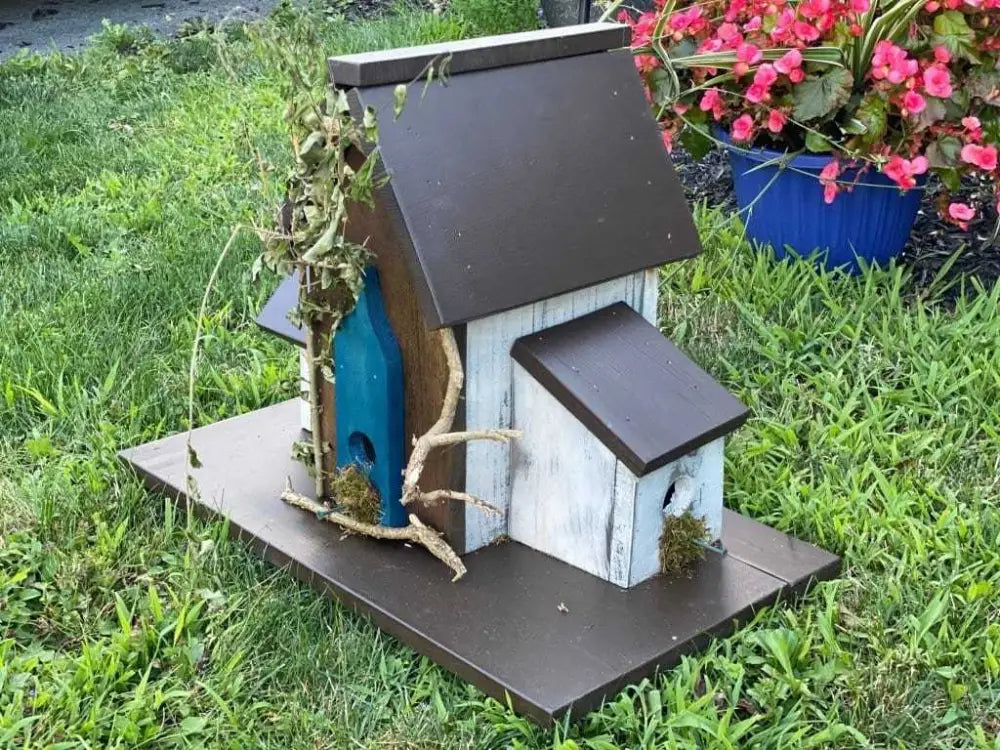 Triple birdhouse with unique design, made from pine wood, featuring three compartments and customizable elements, set in a garden with vibrant flowers.