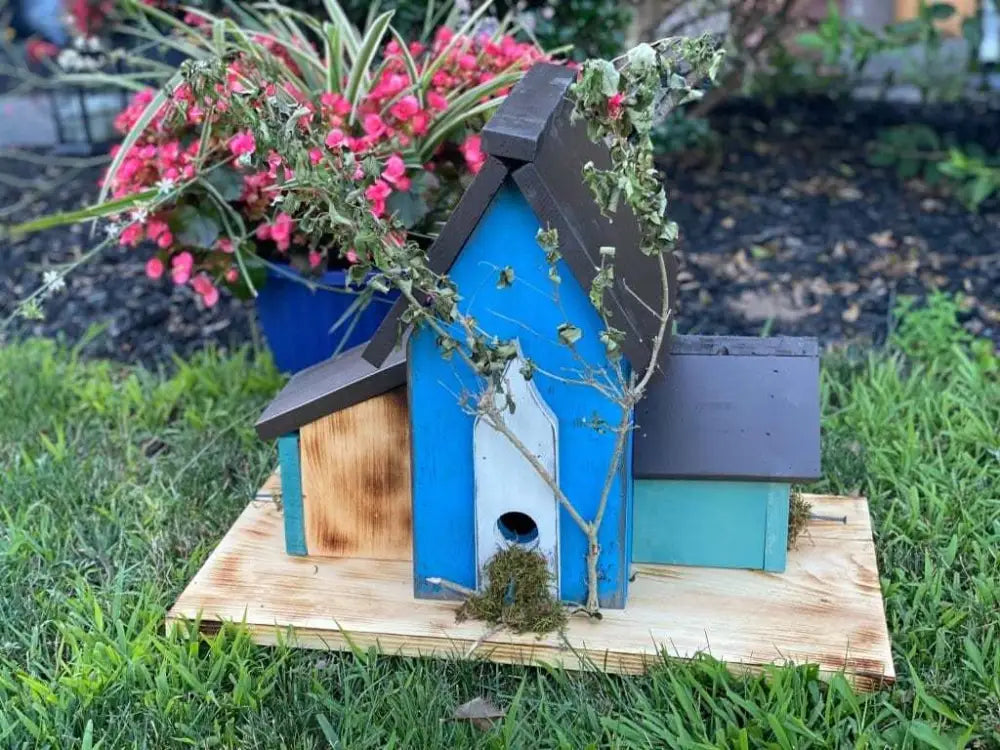 Triple birdhouse made of pine wood with blue and brown tones, featuring three compartments and decorative plants, in a garden setting.