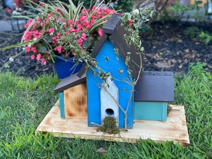 Triple birdhouse made of pine wood with blue and brown tones, featuring three compartments and decorative plants, in a garden setting.