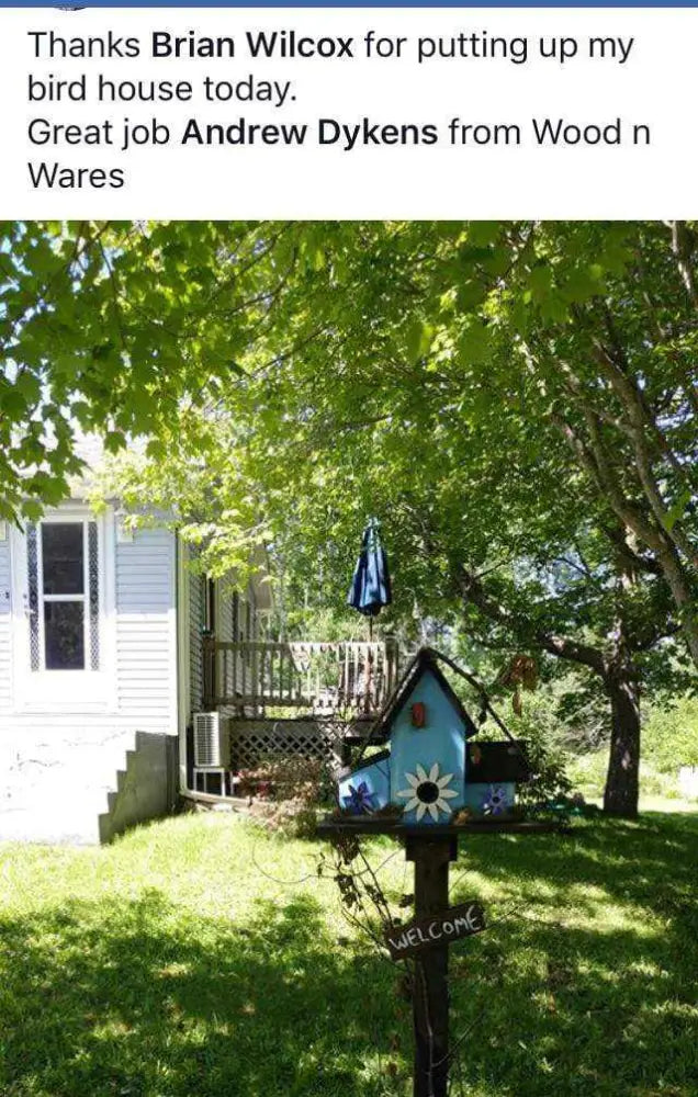 Triple compartment pine wood birdhouse with round entrances and perches, decorated with a sunflower design and welcome sign, mounted on a pole in a green backyard.