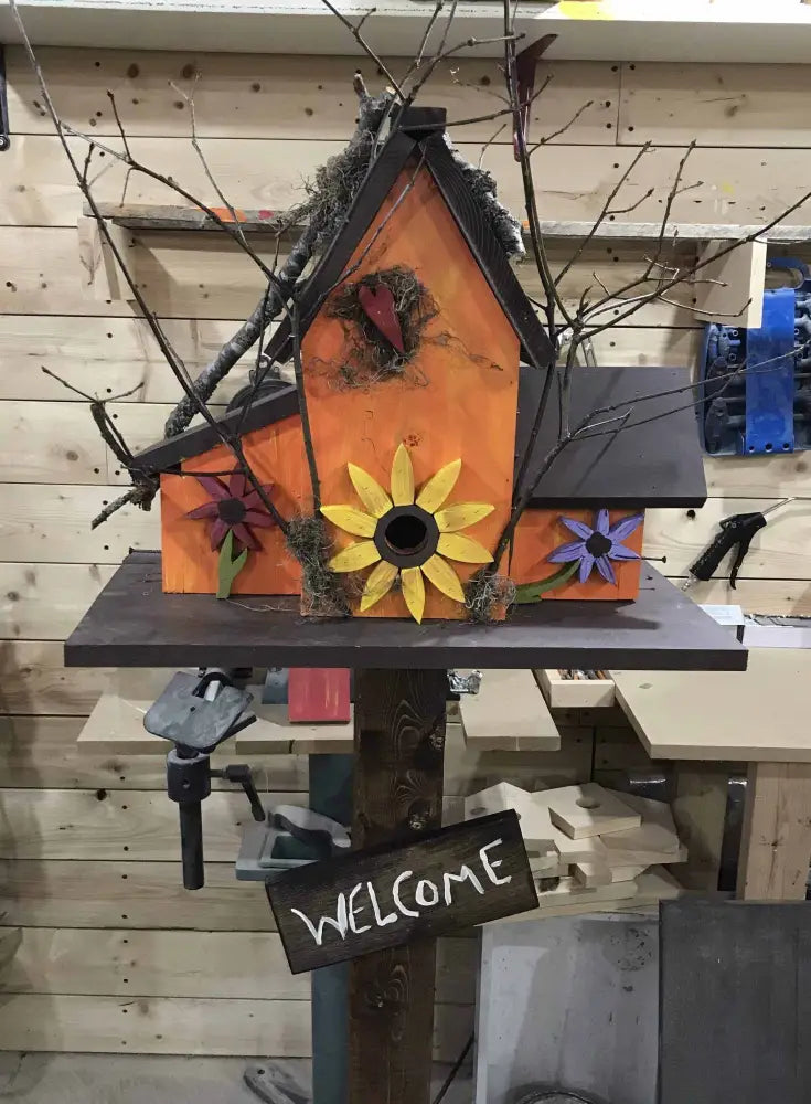 Triple colorful birdhouse with sunflower and flower decorations mounted on wooden post with welcome sign in workshop setting
