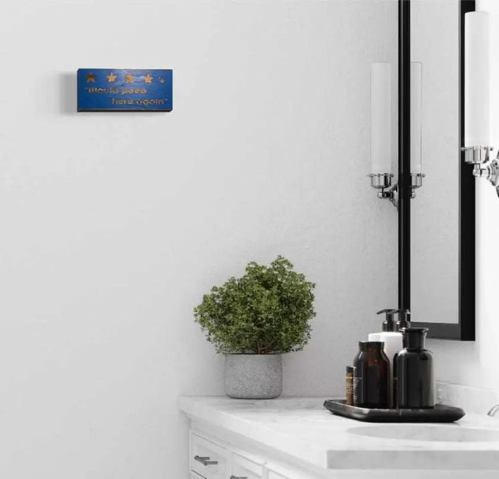 Rustic "Would Poop Here Again" bathroom sign hanging on a white wall above a countertop with a plant.