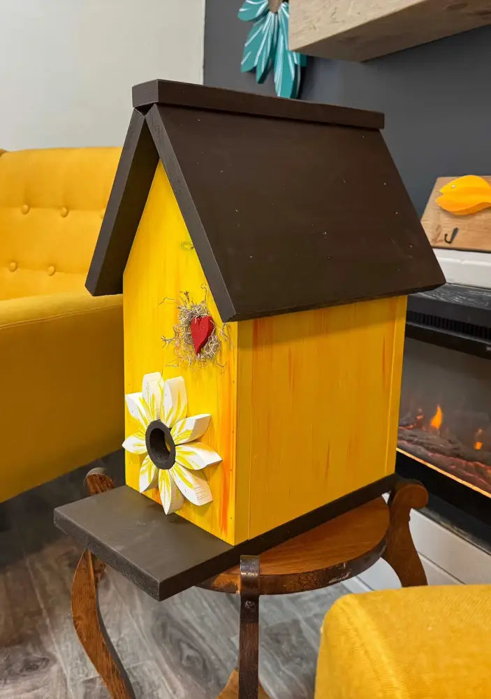 Handcrafted yellow pine birdhouse with dark brown roof and sunflower decoration, indoor setting with yellow furniture and fireplace