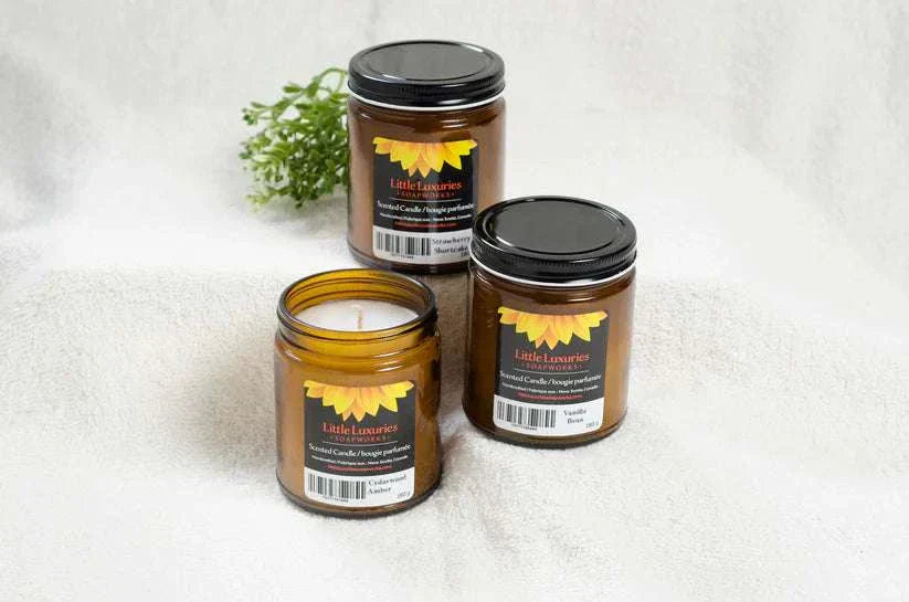 Luxury scented candles in amber glass jars with black lids on a soft fabric background, offering various fragrances for home ambiance.