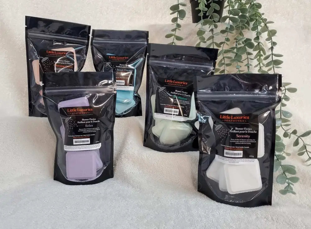 Five black resealable pouches of Little Luxuries shower steamers in various colors displayed on a white towel with green plants in the background