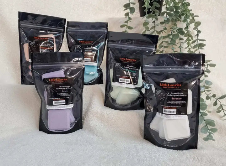 Five black resealable pouches of Little Luxuries shower steamers in various colors displayed on a white towel with green plants in the background