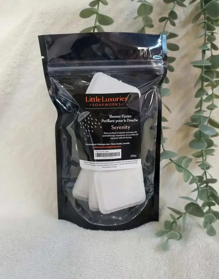 Pack of Little Luxuries Soapworks Serenity Shower Steamers with essential oils in resealable black pouch beside eucalyptus leaves